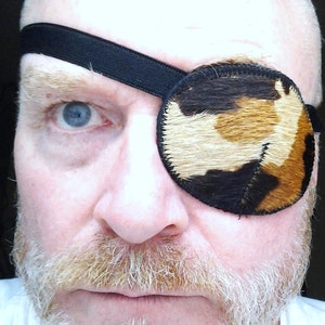 Op de afbeelding: Een man met een witte baard en een zwart ooglapje met een bruin en wit bontpatroon. Het ooglapje wordt op zijn plaats gehouden met een zwarte band.