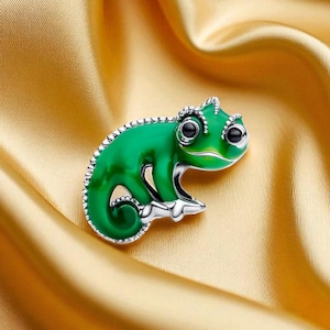 May include: A green chameleon brooch with black eyes and a silver-colored base. The enamel brooch is set against a gold satin background. The brooch is a decorative accessory.