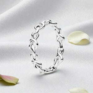 May include: A silver-colored ring with an interwoven knot design. The ring is set against a white, silky background, with scattered flower petals. The ring's intricate pattern is the focal point, showcasing a delicate and elegant aesthetic.