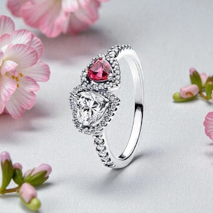 May include: A silver ring featuring two heart-shaped stones, one clear and one pink, each surrounded by small, sparkling accents. The ring's band is also adorned with small, clear stones. Pink flowers are in the background.