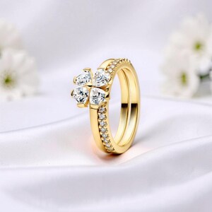Op de afbeelding: Een gouden ringenset. Eén ring heeft een klaverbladontwerp met heldere, sprankelende stenen. De tweede ring is een eenvoudige band met kleine, heldere stenen. De ringen staan tegen een witte achtergrond.