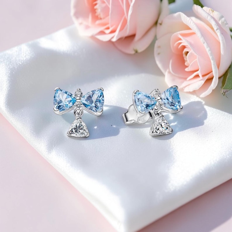 Puede incluir: Pendientes de plata con forma de lazo, con piedras preciosas de color azul claro y un colgante triangular de cristal transparente. Los pendientes se muestran sobre una superficie de sat&eacute;n blanco, con rosas rosas en el fondo.