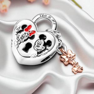 Op de afbeelding: Een zilveren hartvormige hangslotbedel met Mickey en Minnie Mouse. De bedel heeft een sleutelgat en de woorden "YOU & ME" zijn bovenop gegraveerd. Een roségouden sleutelbedel met een strikdesign hangt aan het hangslot.