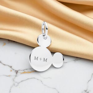 May include: A silver pendant in the shape of a mouse head, with the initials "M + M" engraved on the front. The pendant has a loop at the top for a chain and is displayed on a gold satin fabric.