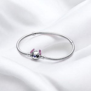 Puede incluir: Una pulsera plateada con un charm de Stitch. El charm presenta al personaje de Lilo & Stitch, con orejas rosas y detalles azules. La pulsera tiene un diseño de cadena de serpiente, con una circunferencia de unos 18 cm.