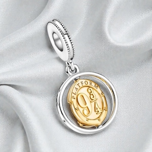 May include: A silver charm with a gold-colored oval pendant featuring the text "PLATFORM 9 3/4". The charm has a circular design with a beaded edge and a loop for attaching to a bracelet or necklace. The background is a white, silky fabric.