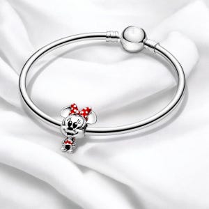 Pu&ograve; includere: Un braccialetto con ciondolo in argento con un ciondolo in argento di Minnie Mouse. Minnie Mouse indossa un vestito rosso con pois bianchi e un fiocco rosso.