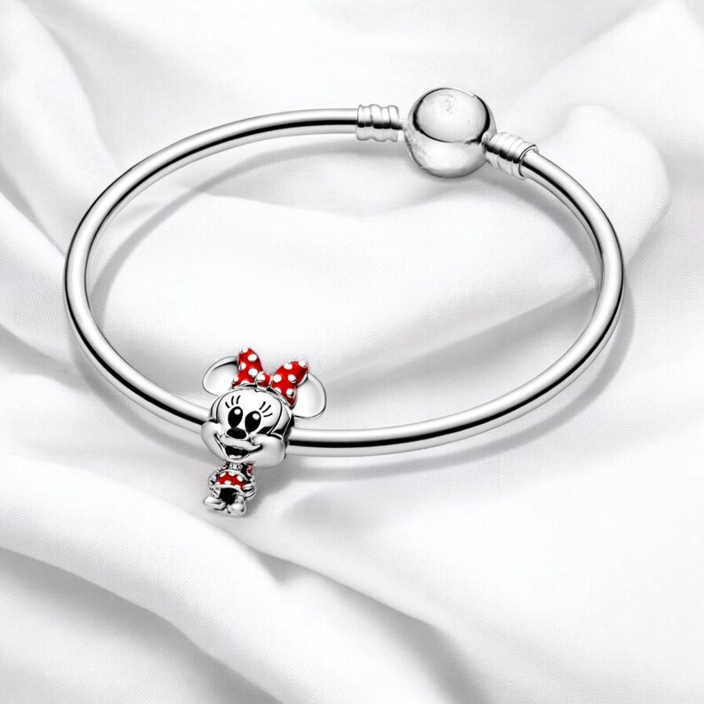 Pu&ograve; includere: Un braccialetto con ciondolo in argento con un ciondolo in argento di Minnie Mouse. Minnie Mouse indossa un vestito rosso con pois bianchi e un fiocco rosso.