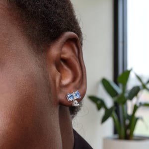 Puede incluir: Primer plano de una oreja con pendientes de plata. Cada pendiente presenta dos piedras azules claras en forma de coraz&oacute;n y una piedra m&aacute;s peque&ntilde;a y transparente. Los pendientes est&aacute;n colocados contra la piel y el cabello de la persona.