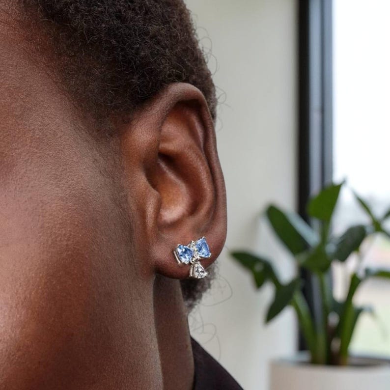 Puede incluir: Primer plano de una oreja con pendientes de plata. Cada pendiente presenta dos piedras azules claras en forma de coraz&oacute;n y una piedra m&aacute;s peque&ntilde;a y transparente. Los pendientes est&aacute;n colocados contra la piel y el cabello de la persona.
