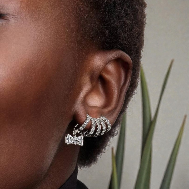 May include: Close-up of an ear displaying two sets of earrings. One set has a bow-shaped charm, the other multiple stacked hoops, all with sparkling stones. The earrings are silver-toned.