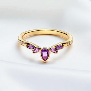 May include: A gold-colored ring with a curved design, featuring a central pear-shaped purple gemstone and smaller purple stones on each side. The band is adorned with small, clear stones. The ring is set against a white background.