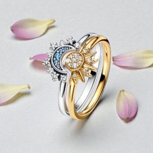 Puede incluir: Un conjunto de dos anillos, uno plateado y otro dorado, con diseños celestiales. El anillo plateado tiene una luna creciente con piedras azules y transparentes. El anillo dorado tiene un diseño de sol con piedras transparentes. Pétalos de flores rosas y amarillas.