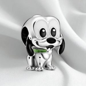 Könnte beinhalten: Ein silberfarbener Charm in Form eines Cartoon-Hundes mit schwarzen Ohren und Nase. Der Hund hat große, runde schwarze Augen und ein grünes Halsband. Der Charm ist vor einem weißen Hintergrund platziert.