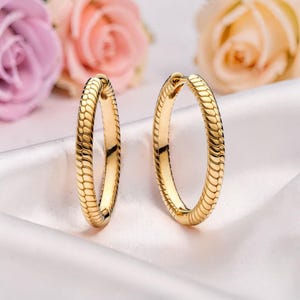 May include: A pair of gold-colored hoop earrings. The earrings have a textured, snake-like pattern. The background features soft pink and peach-colored roses on a white satin surface.