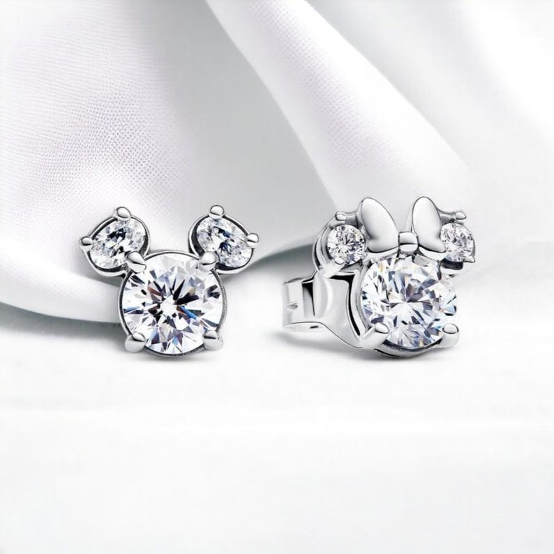 May include: A pair of silver earrings with a clear crystal in the center, shaped like Mickey Mouse and Minnie Mouse heads.