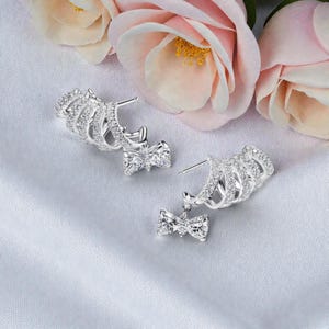 May include: Silver earrings with a bow design. The earrings have a curved, openwork design with embedded clear crystals. Each earring has a small bow with a central crystal. Pink flowers are in the background.