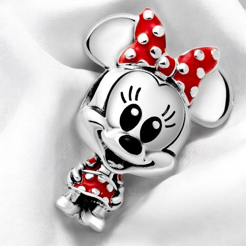 Pu&ograve; includere: Un ciondolo d'argento con un'immagine di Minnie Mouse in stile cartone animato. Indossa un vestito rosso con pois bianchi e un fiocco rosso con pois bianchi.