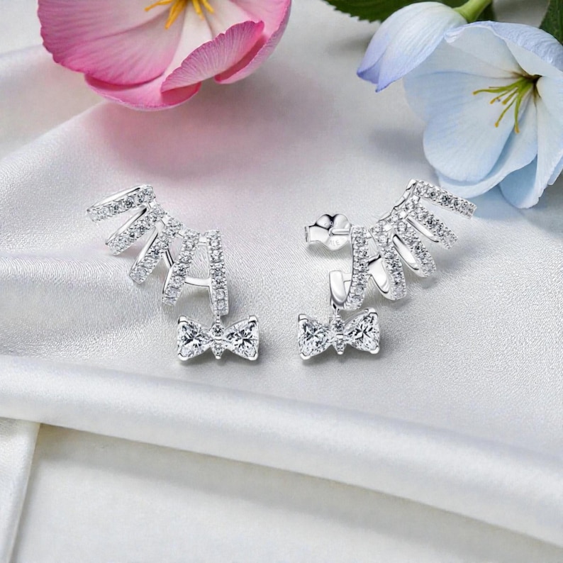 May include: A pair of silver earrings with a bow design. The earrings feature a cluster of small, sparkling stones and a bow-shaped pendant. The earrings are displayed on a white satin surface, with pink and blue flowers in the background.