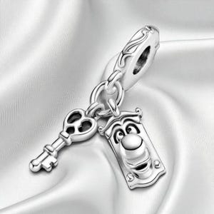 May include: A silver charm with a key and a door knocker design. The key has a heart-shaped cutout, and the door knocker has a smiling face. The charm is attached to a silver loop, suitable for a bracelet or necklace.