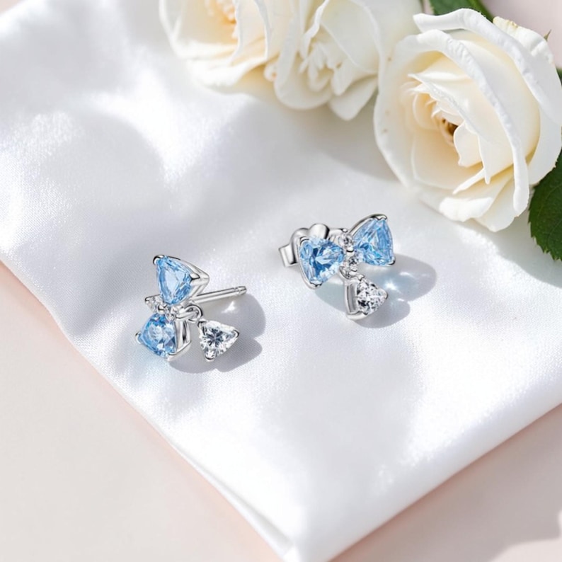 Puede incluir: Pendientes de plata con dise&ntilde;o de lazo adornados con piedras preciosas azul claro y transparentes. Los pendientes est&aacute;n colocados sobre una tela de sat&eacute;n blanco, con rosas blancas en el fondo.