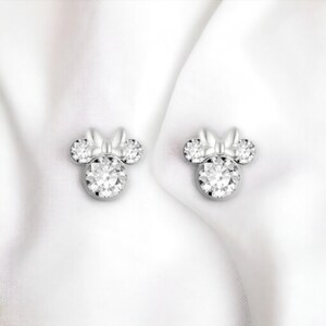May include: A pair of silver earrings with a clear crystal design in the shape of a cartoon mouse with a bow.