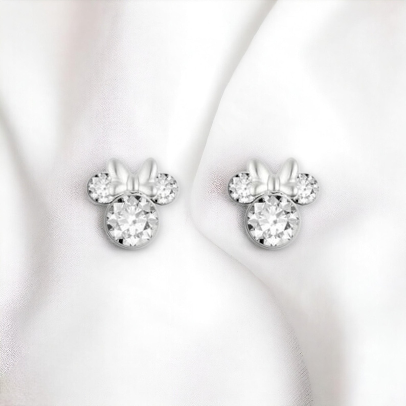 May include: A pair of silver earrings with a clear crystal design in the shape of a cartoon mouse with a bow.