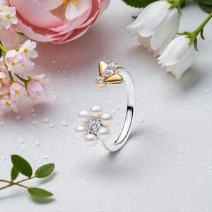 Könnte beinhalten: Ein silberner Ring mit floralem Design. Der Ring hat eine Ansammlung weißer Perlen und einen zentralen diamantartigen Stein. Die Oberseite des Rings hat ein goldfarbenes Schmetterlingsdesign mit einem diamantartigen Stein. Der Ring ist vor einem weißen Hintergrund mit Blumen platziert.