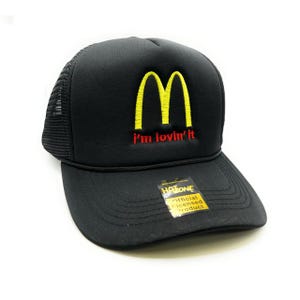 Puede incluir: Gorra de camionero negra con un logotipo "M" amarillo y el texto "i'm lovin' it" en rojo. La gorra es un producto con licencia oficial.