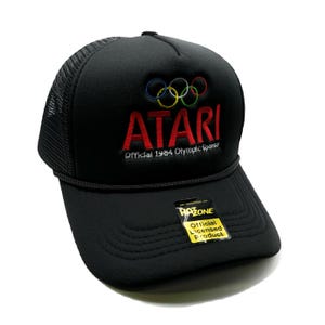 May include: Black mesh trucker hat with a red "ATARI" logo and the Olympic rings in colorful embroidery. The text "Official 1984 Olympic Sponsor" is below the logo.