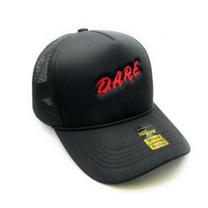 Peut inclure: Casquette de camionneur noire avec un logo "DARE" brodé en rouge. La casquette a un dos en mesh et une sangle réglable noire avec une étiquette jaune "Produit officiel sous licence".