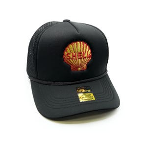 Könnte beinhalten: Schwarze Trucker-Kappe mit einem goldenen und roten Shell-Logo, das auf der Vorderseite gestickt ist. Die Kappe hat ein schwarzes Netzrücken und ein goldenes Etikett mit der Aufschrift "Offizielles Lizenzprodukt" an der Seite.