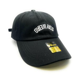 Può includere: Un cappello da baseball nero con un testo bianco ricamato ad arco che dice "Stay Weird". Il cappello ha un'etichetta gialla che dice "Prodotto ufficiale con licenza" e "MAZONE".