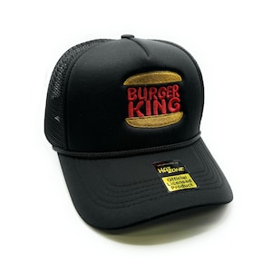 May include: Black trucker hat with a Burger King logo embroidered on the front. The logo is red and yellow with a burger graphic. The hat has a black mesh back and a black brim. The hat has a yellow tag that says "Official Licensed Product" on the side.