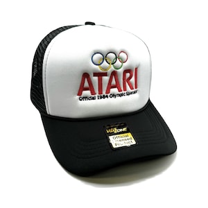 May include: A black and white trucker hat with a white front panel featuring the Atari logo and the Olympic rings. The text "Official 1984 Olympic Sponsor" is embroidered below the logo. The back of the hat is black mesh. A yellow tag with the text "HA ZONE Official Licensed Product" is attached to the hat.