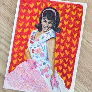 Könnte beinhalten: Eine Frau mit dunklen Haaren und einem rosafarbenen Stirnband trägt ein weißes Kleid mit rosa und floralen Stickereien. Sie steht vor einem roten Hintergrund mit orangenen Herzen.