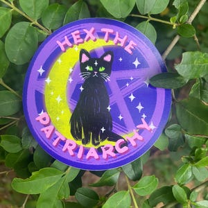 May include: A round sticker with a black cat illustration, green eyes, and the text "HEX THE PATRIARCHY". The design features a purple background with a crescent moon, stars, and a pentagram.