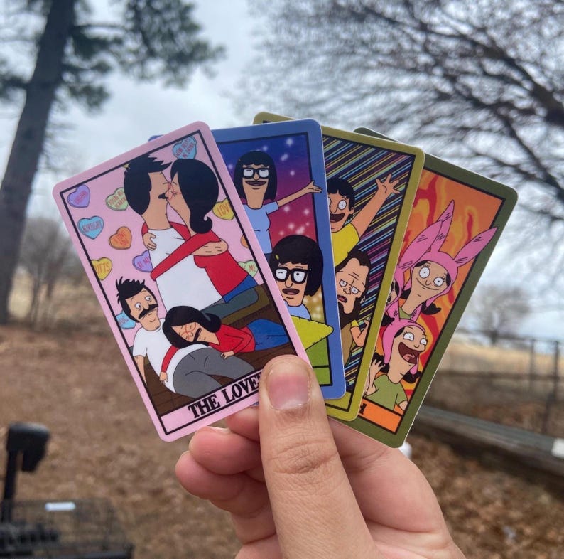May include: Four colorful cards featuring cartoon characters. The cards have different backgrounds and character arrangements. The first card has the text "THE LOVE" and a couple kissing. The other cards feature other characters. The cards are held in a hand.