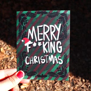 May include: A Christmas card with the text "MERRY F***KING CHRISTMAS" in white, with a Santa hat on the letter F. The card has a green and burgundy striped background with a black leaf border.