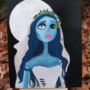 May include: A painting of a blue-skinned bride with long blue hair, wearing a white dress and veil, set against a black background with a white moon. A green worm is on her head. The artwork is displayed on a small wooden easel.