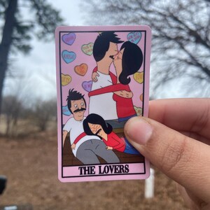 May include: A pink sticker featuring a cartoon image of a family from the animated series, Bob's Burgers. The sticker depicts two couples, one kissing and the other embracing. The text "THE LOVERS" is at the bottom.
