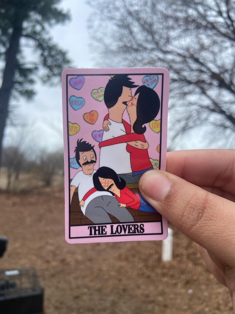 May include: A pink sticker featuring a cartoon image of a family from the animated series, Bob's Burgers. The sticker depicts two couples, one kissing and the other embracing. The text "THE LOVERS" is at the bottom.