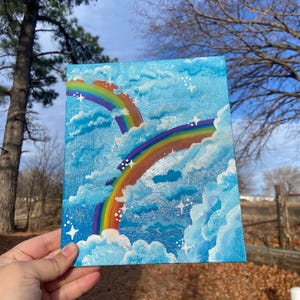 Pu&ograve; includere: Un piccolo dipinto quadrato con due vivaci arcobaleni che si estendono su un cielo blu brillante pieno di soffici nuvole bianche e stelle scintillanti. L'opera d'arte &egrave; tenuta in mano, mostrando la pittura acrilica.