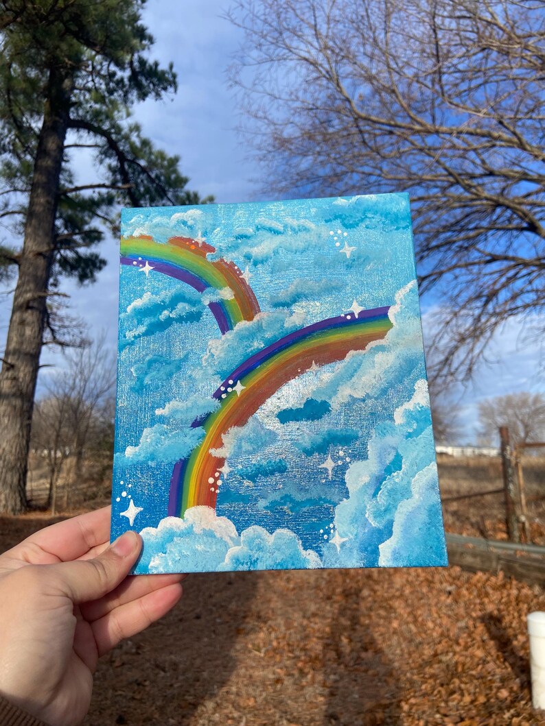 Pu&ograve; includere: Un piccolo dipinto quadrato con due vivaci arcobaleni che si estendono su un cielo blu brillante pieno di soffici nuvole bianche e stelle scintillanti. L'opera d'arte &egrave; tenuta in mano, mostrando la pittura acrilica.