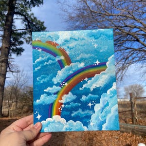 May include: A small, square painting shows two rainbows arcing across a blue sky with fluffy white clouds and sparkling stars. The rainbows display a full spectrum of colours, adding vibrancy to the artwork.