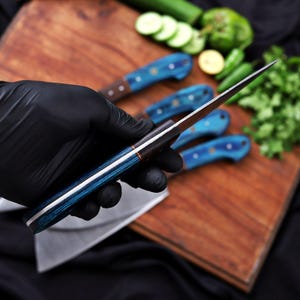 May include: A close-up of a knife with a blue and white handle, held by a gloved hand. The knife blade is silver and sharp. Other knives with blue handles are on a wooden cutting board. Fresh vegetables are in the background.