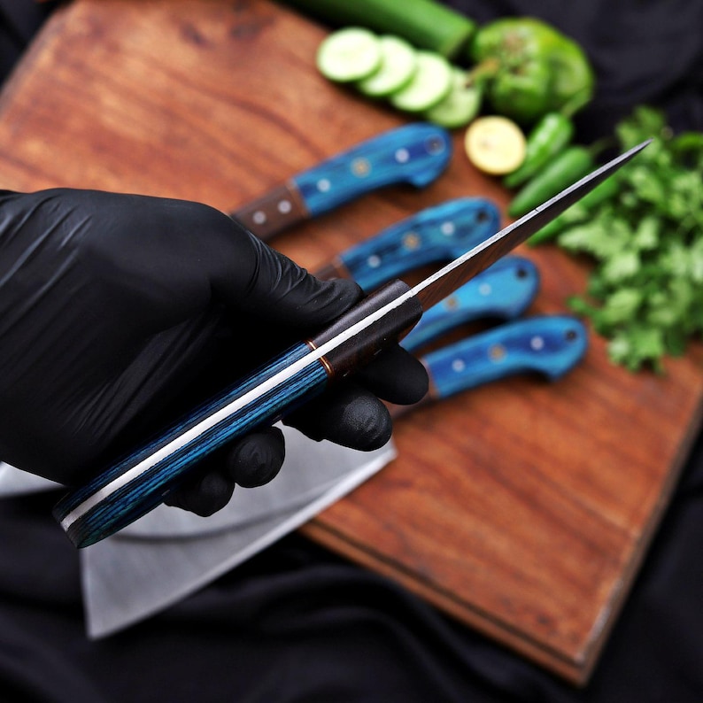 May include: A close-up of a knife with a blue and white handle, held by a gloved hand. The knife blade is silver and sharp. Other knives with blue handles are on a wooden cutting board. Fresh vegetables are in the background.