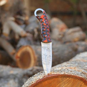May include: A handcrafted knife with a patterned blade and a handle featuring a red and black design. The knife has a ring at the end of the handle. The blade is made of steel and has a unique pattern.