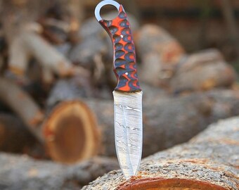 May include: A handcrafted knife with a patterned blade and a handle featuring a red and black design. The knife has a ring at the end of the handle. The blade is made of steel and has a unique pattern.