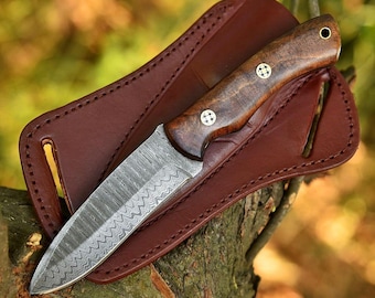 May include: A Damascus steel knife with a wooden handle and a brown leather sheath. The knife blade has a unique wave pattern. The handle is secured with three decorative rivets. The sheath is stitched and has a belt loop.